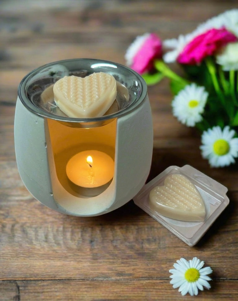 Heart-shaped wax melts in a small wax warmer on a wooden surface with flowers in the background. Amor Scent Co. Fragrance scent Aroma